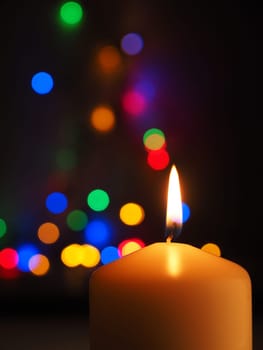 A white candle is burning with colorful Christmas lights bokeh in the background, space for text