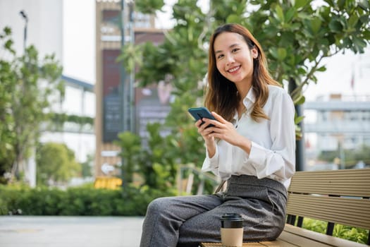Asian young woman sitting on bench outdoor on park using smart mobile phone, Smiling businesswoman using smartphone outside office, lifestyle portrait pretty texting message, coffee paper cup takeaway