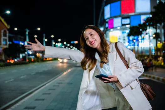 Asian businesswoman standing hail waving hand taxi on road in busy city street at night, beautiful woman smiling using mobile phone application hailing with hand up calling cab after late work
