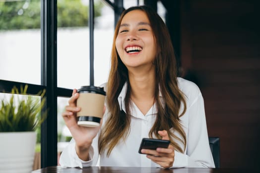 Asian young business woman holding coffee paper cup and using mobile phone to online chating at coffee shop, Happy female sitting in cafe near window she holding mobile phone on hand