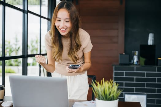 Excited female smile got good news on smartphone while working on computer at cefa, social media network, Asia young woman holding mobile smart phone raising hands celebrate with laptop, happy success