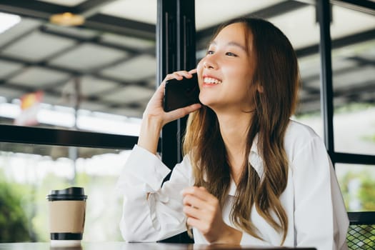 Beautiful young businesswoman freelance lunch outside holding smartphone and coffee paper cup at coffee shop, Happy female lifestyle sitting in cafe near window she using mobile phone