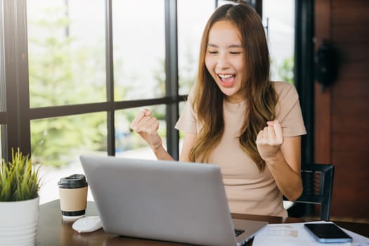 Excited happy Asian business young woman sitting with laptop computer at cafe desk raising hands up and celebrating success, female feeling winner rejoicing online win got new job at home