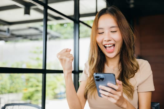Asian young woman celebrating work success near windows in morning with mobile phone, Beautiful female successful excited she saised hands with smartphone winner gesture outside