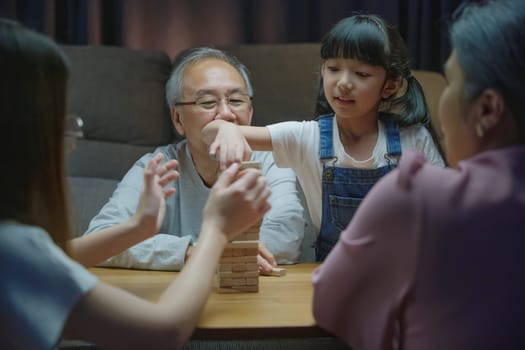 Happy grandparents Asian family enjoy playing toy block with little daughter and mother together at home night time, Smiling parent having fun play build constructor tower of wooden blocks, education