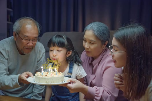 Happy Asian family grandparents and mother celebrating enjoy birthday party with cake of granddaughter Blowing candle at home in night time, Birthday surprise concept