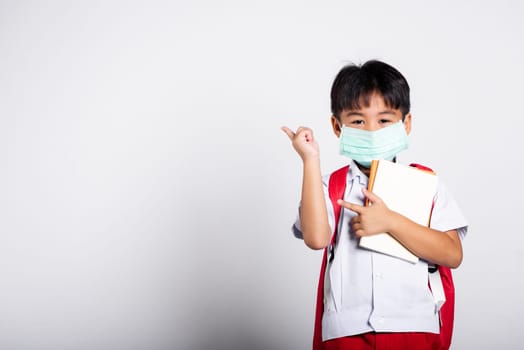 Asian student kid boy wear student thai uniform and protect face mask ready to go to school pointing finger to space in studio shot isolated on white background, preschool, new normal back to school