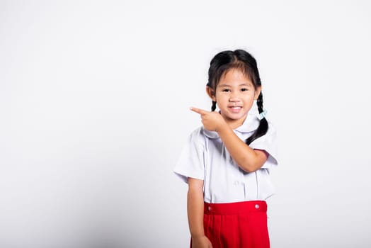 Asian toddler smiling happy wear student thai uniform red skirt keeps pointing finger at copy space in studio shot isolated on white background, Portrait little children girl preschool, Back to school