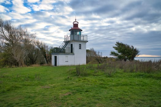 Lighthouse, Spodsbjerg Fyr in Huntsted on the coast of Denmark. Sun rays shining through the clouds. Meadow with trees. Landscape photo from sea