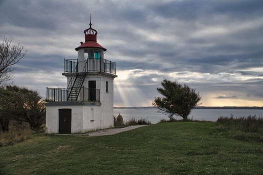Lighthouse, Spodsbjerg Fyr in Huntsted on the coast of Denmark. Sun rays shining through the clouds. Meadow with trees. Landscape photo from sea