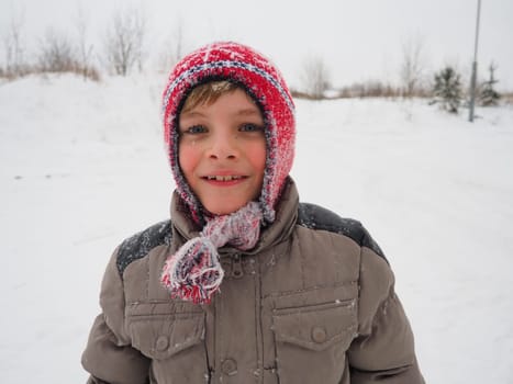 A cute boy 8 years old in a knitted red hat with braids and tassels. The child smiles cheerfully. Snowflakes on the face. The theme of winter holidays, frosty weather and travel in the north
