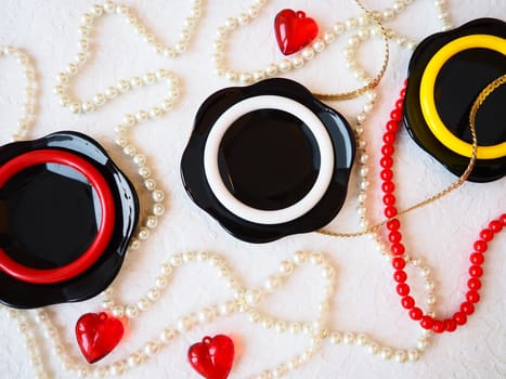 Flat lay. A beautiful composition of glass black dishes, plastic jewelry, bracelets, beads, red hearts on a white background. Women's baubles for February 14, Valentine's Day. Still life.