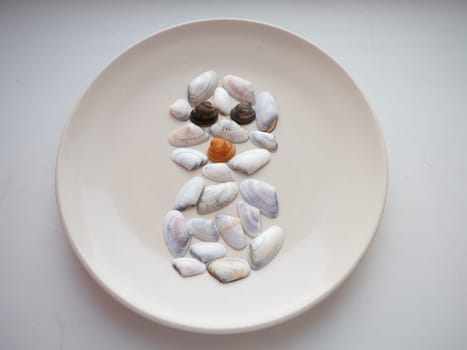 Cute snowman made of seashells. The seashells are laid out on a white plate in the shape of a snowman with a nose and eyes. Pastel colors, soft focus. White background.