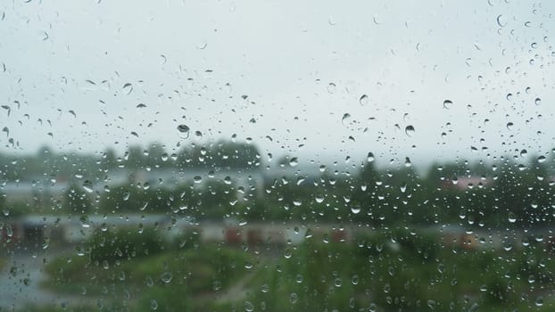 Raindrops on the glass window on a rainy day. Water drips from above. Blurred background. Street with burnings and trees outside the window. It's raining in the summer
