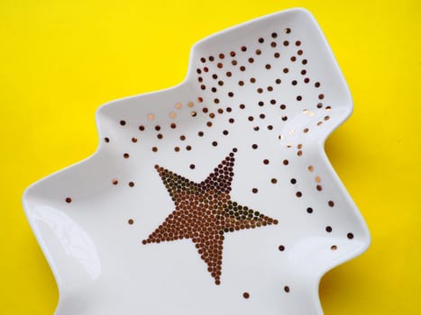White plate in the shape of a Christmas tree with gold decorations and a star in the middle. Yellow background. New Year or Christmas card. Background for advertising and congratulations. Copy space.