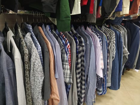 Clothes hung on hangers. Assortment of second hand store. Men's shirts hanging on a rack, ready for sale to customers. Saving money in crisis. Cheap shopping. Checkered, striped, plain men's shirts.