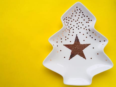 White plate in the shape of a Christmas tree with gold decorations and a star in the middle. Yellow background. New Year or Christmas card. Background for advertising and congratulations. Copy space.