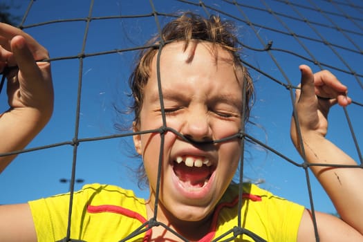 An 8-year-old Caucasian girl stands behind a sports net and screams. Volleyball sports dividing net against the blue sky close-up. Outdoor sports