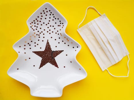 White plate in the shape of a Christmas tree with gold decorations and a star in the middle. Yellow background. New Year or Christmas card. White medical mask. Covid holidays tainted by lockdown