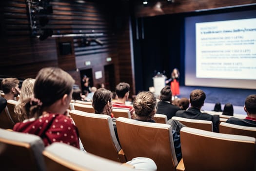Business and entrepreneurship symposium. Female speaker giving a talk at business meeting. Audience in conference hall. Rear view of unrecognized participant in audience.