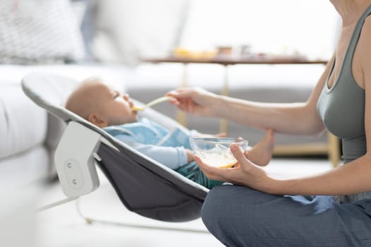 Mother spoon feeding her baby boy child in baby chair with fruit puree. Baby solid food introduction concept