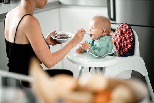 Mother spoon feeding her baby boy child in baby chair with fruit puree in kitchen. Baby solid food introduction concept.
