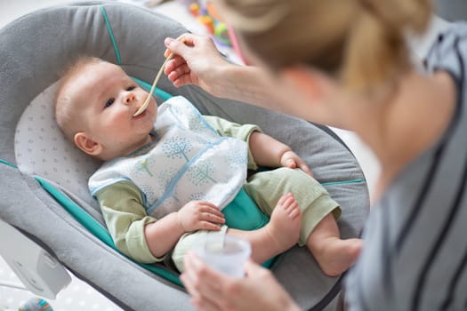 Mother spoon feeding her baby boy child in baby chair with fruit puree. Baby solid food introduction concept