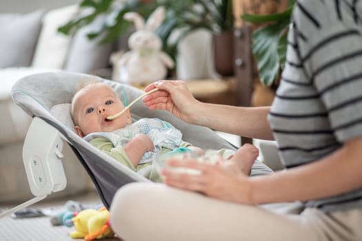 Mother spoon feeding her baby boy child in baby chair with fruit puree. Baby solid food introduction concept