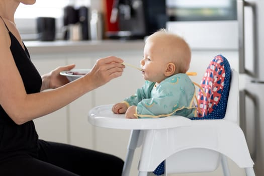 Mother spoon feeding her baby boy child in baby chair with fruit puree in kitchen at home. Baby solid food introduction concept.