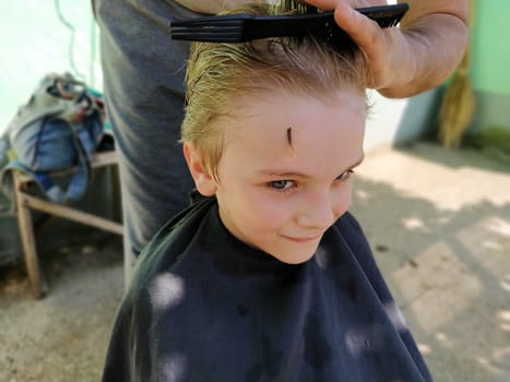 The hairdresser cuts the boy. The child is smiling and looking at the camera. Toddler 7 years old wants a beautiful hairstyle. A lock of hair fell over his forehead. Hands of the hairdresser.