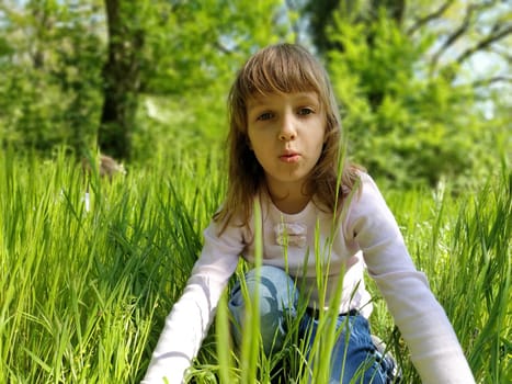 Nice girl in the green grass. The child has blond long hair and a white blouse. The girl looks at the camera and spreads tall green grass with her hands. The concept of ecology and harmony with nature.