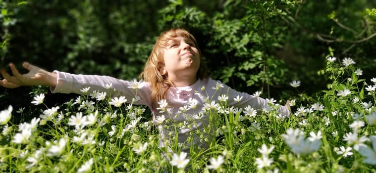 Cute girl with blond hair in the meadow. Wildflowers are white. Stellaria is a genus of flowering plants in the clove family. The baby is happy. The girl spread her arms and looks up and smiles