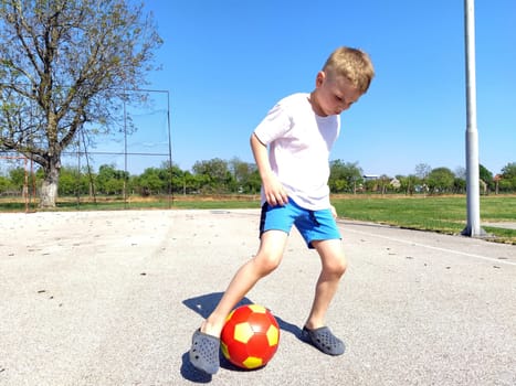 Sremska Mitrovica, Serbia. June 6, 2020. A boy plays ball at the playground. Asphalt sports court. A child in a white t-shirt. Toddler with blond hair, blond, 7 years old. Running, kicking and endurance exercises. Sunny weather and blue sky in the background