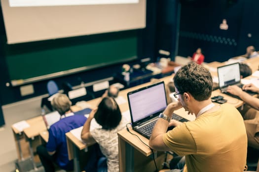 Workshop at university. Rear view of students sitting and listening in lecture hall doing practical tasks on their laptops.