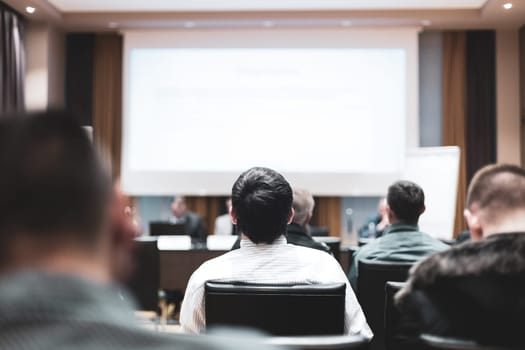 Business and entrepreneurship symposium. Speaker giving a talk at business meeting. Audience in the conference hall. Rear view of unrecognized participant in audience.