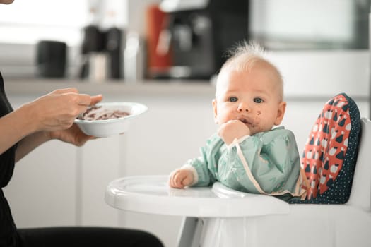 Mother spoon feeding her baby boy child in baby chair with fruit puree in kitchen at home. Baby solid food introduction concept