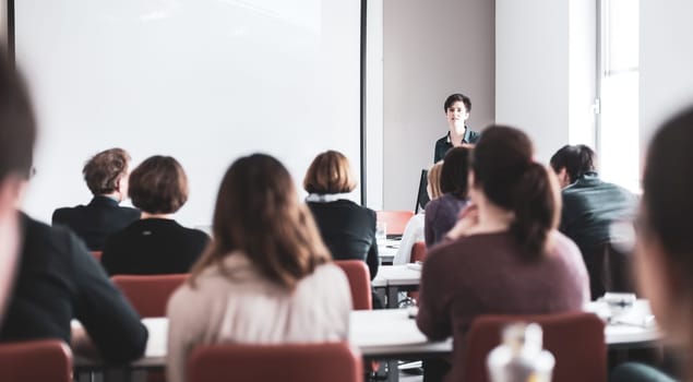 Female speaker giving presentation in lecture hall at university workshop . Participants listening to lecture and making notes. Scientific conference event.