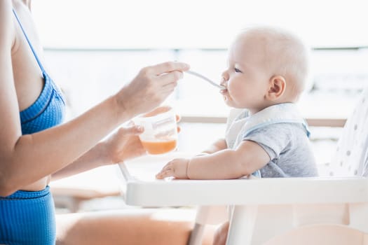 Mother spoon feeding her baby boy child in baby chair with fruit puree on a porch on summer vacations. Baby solid food introduction concept