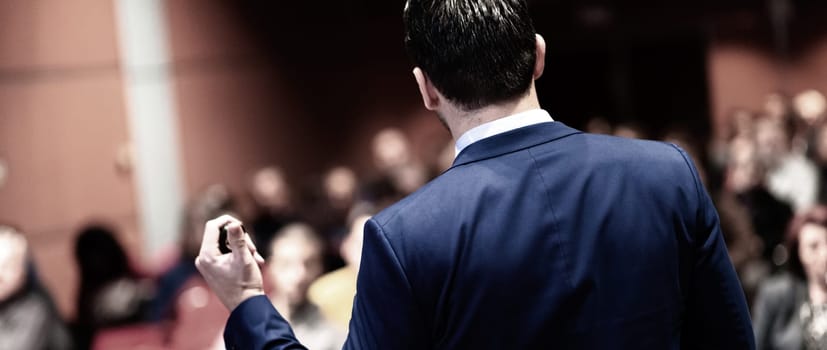 Rear view of speaker giving a talk on corporate Business Conference. Audience at the conference hall. Business and Entrepreneurship event. Panoramic composition.