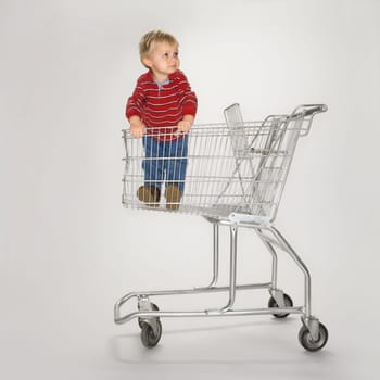 Studio portrait of Caucasian boy standing alone in empty shopping cart against white background.