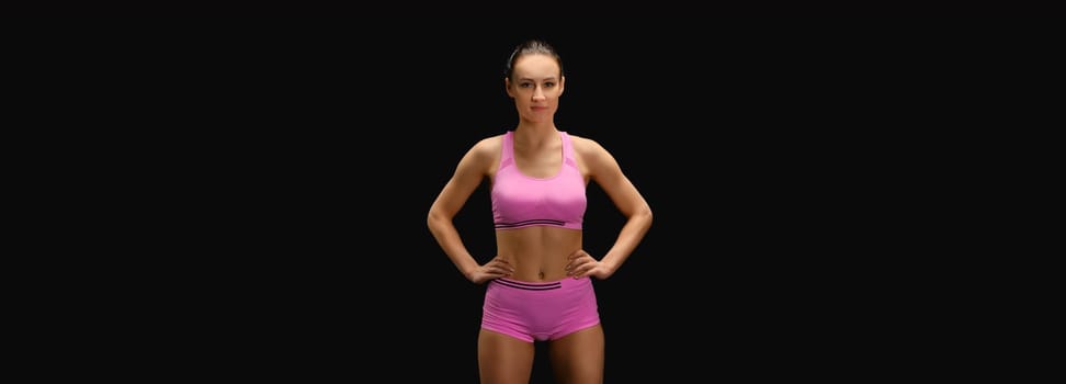 Athletic young woman in pink sportswear with hands on waist over black background