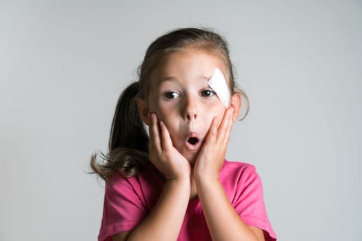 Close up surprised, cute, nice dark haired little girl with bandage patch on face, wear pink shirt on white background. Afraid of parents reaction. Physical wounds, fidget children, doctor fear