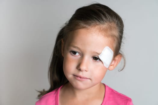 Close up sad, nice, lovely, adorable dark haired little child, girl with white patch bandage on face, looking down. Child abuse and physical wounds. Kid protection and safety, healthcare and medicine