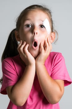 Close up funny, adorable dark haired little girl with injury and patch on face, wear pink shirt on white background. Afraid of parents reaction. Physical wounds, fidget children and active lifestyle