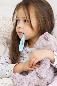 Vertical sad, calm. depressed, tired little child, girl looking away and holding electronic thermometer in mouth, embracing teddy bear in bed. Seasonal cold, influenza and virus infection disease