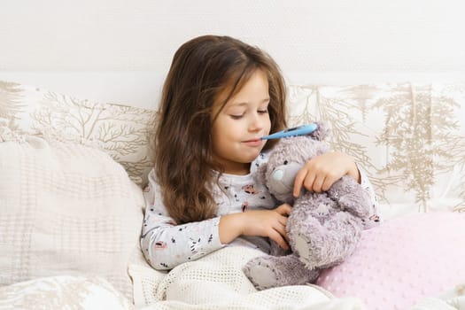 Smiling, confident, positive little child, girl holding electronic thermometer in mouth, hugging teddy bear and calming toy in bed. Covid 19, seasonal cold, virus infection disease. Copy space