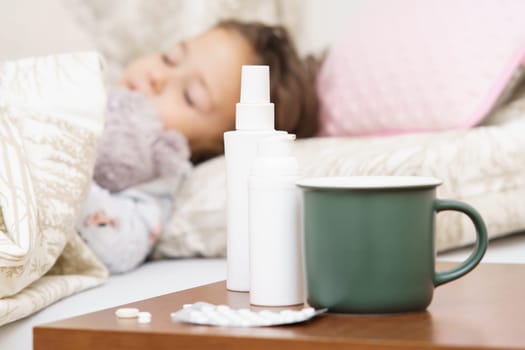 Close up green mug, nasal and throat sprays containers and pills blister on bed table against defocused sick little child, girl sleeping in bed. Pharmacy and remedy as painkillers, antibiotics.
