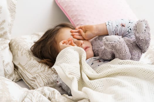 Sad, disappointed, crying child girl rubbing eyes, holding stuffed toy, lying in soft white bed with pillows in the morning. Hurt and family child abuse, hard relationship. Social issues, loneliness