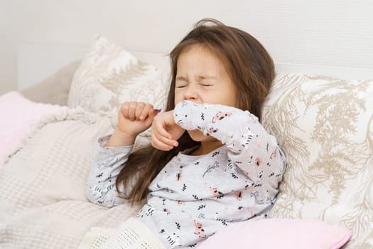 Exhausted little long haired brunette girl yawning or sneezing with close eyes, covering mouth with hand, wearing cute pajamas and lying in white bed. Broken and not deep sleep, kid nightmares