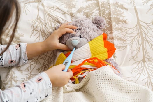 Cropped rearview of little child, girl healing bear toy, checking temperature with electronic thermometer in toy mouth, wrapping in scarf on bed, sit on bed in bedroom. Little doctor help friend